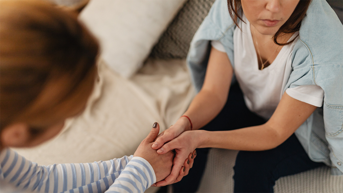 Two women holding hands, one sitting on sofa, looking away and the other standing looking down towards her in a caring manner.